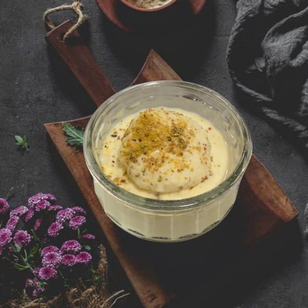 A glass bowl of Rasmalai, an Indian dessert, with crushed nuts on a wooden board.
