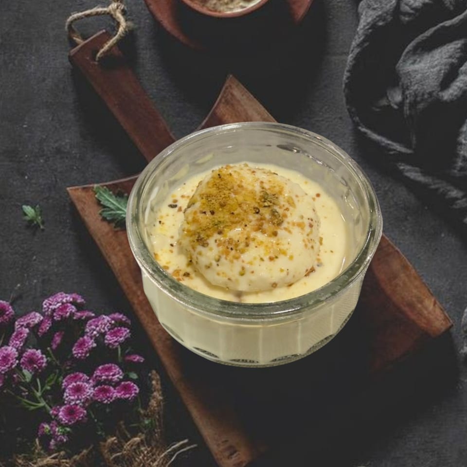 A glass bowl of Rasmalai, an Indian dessert, with crushed nuts on a wooden board.