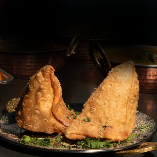 Two golden-brown samosas garnished with fresh herbs on a dark plate, with copper pots in the background.