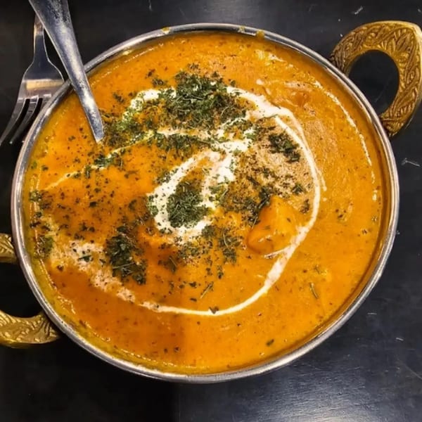 A close-up of a vibrant orange Afghan curry with cream swirls and green herbs in a metal bowl.